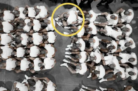 An image of prisoners sitting on a floor in white socks and white shorts and T-shirts. Their shaved heads are tilted so we do not see their faces. A yellow circle indicates Mr. Garcia, and his hand tattoos