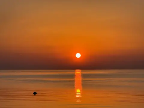 William Dickson A sunset over water, with the sun casting an orange glow over the water.