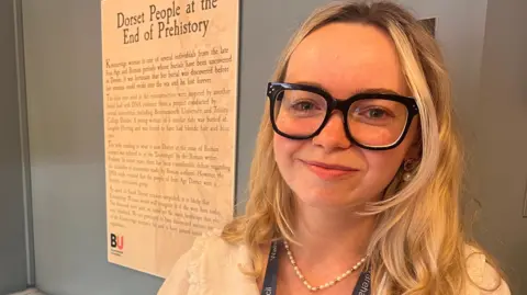 Katie Seal smiles at the camera. She has long blonde hair and is wearing large black-framed glasses with very thick rims.