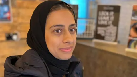 Mariam smiles as she stands in the cafe in a black headscarf and coat.