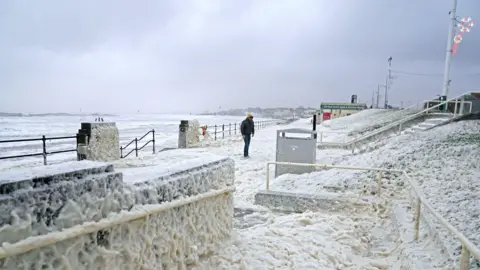 PA Media A man walks through sea foam in Seaburn, Sunderland, as Storm Babet batters the country.