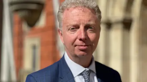 BBC Alex Allinson is wearing a navy suit, light blue shirt and spotted tie. He's standing in front of a brick building and has short fair hair.