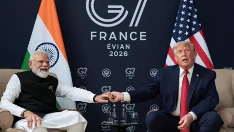 Modi, wearing white and black, holds the hand of Donald Trump, who is wearing a suit and red tie. They sit on chairs in front of an Indian and a US flag, with a 'G7 Evian 2026' sign in the background