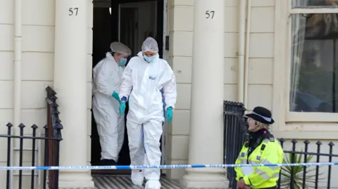 Eddie Mitchell Forensic police officers leave a cream building, guarded by a uniformed police officer and police tape.