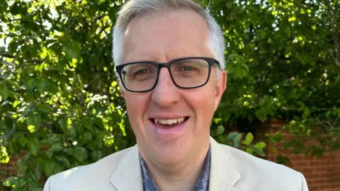 University of Reading A man with grey hair and wearing a white blazer and black glasses, smiling at the camera. There's a brick wall and a large bush behind him.