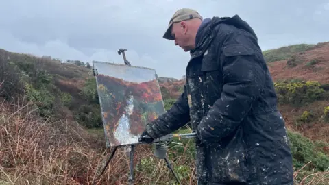 An artist wearing a thick black coat splashed with paint. Leans across to paint on a tripod set up above the banks of a river. The artwork reflects the dark reds and greens of the plant life that flanks the river banks. 
