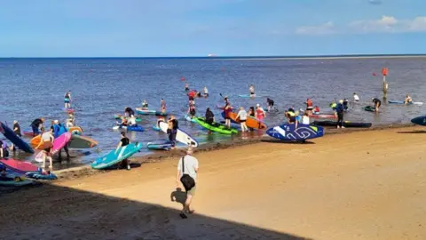A group of paddleboarders on the sand walking into the sea with their boards that are all different colours. Another man wearing shorts and a white top and black bag walks by.
