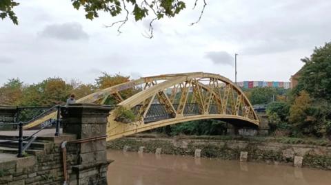 Langton Street Bridge, commonly known as the Banana Bridge, is in the centre of the image. It is a bright yellow curved bridge, with lots of graffiti visible. The wall beneath the bridge, made up of bricks, is shown at high tide, and appears to be very uneven with bricks missing. 