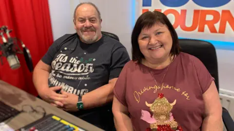 A man and woman are sitting side by side smiling at the camera. The man has grey hair and a grey beard and is wearing a grey Christmas t-shirt. The woman has chin length brown hair and is wearing a plum coloured Christmas t-shirt. They are both sitting behind radio broadcasting equipment.