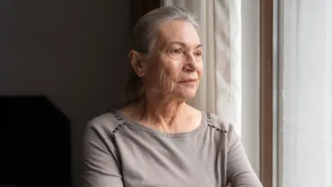 An older woman with long silver hair looks out a bedroom window. She wears a pale grey top with silver beads around the shoulders.