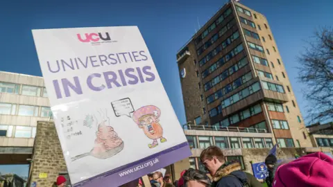 Protesters outside a university building hold a UCU placard reading “Universities in Crisis” during a daytime demonstration.