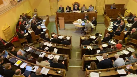 BBC A photo looking down on a council chamber. People are sitting in curved rows with paperwork and computers in front of them.
