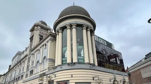 Exterior of Bradford's Alhambra Theatre. A grey and white stone building, with several cupolas and columns supporting them, with a glass block to the right, topped with a green sign saying "Wicked" under a cloudy sky.
