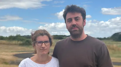 BBC A couple in their thirties standing in front of a grassy site, both looking disappointed. The man is wearing a brown t-shirt while the woman is wearing a white top