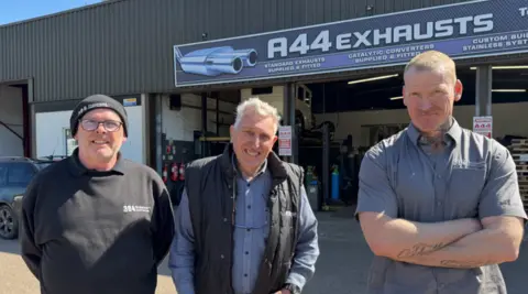Three men are looking at the camera - the man on the left is the only one wearing glasses and a black hat. The A44 Exhausts facility, including a sign with its name, is behind them.