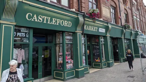 A market building with a green and gold front displays large “CASTLEFORD MARKET” lettering. People walk past shop windows showing sweets, pet supplies and clothing.