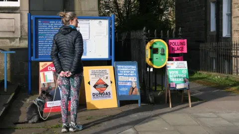 A woman standing outside a polling station with her dog. There are a number of posters for various parties on display on boards. The woman has her back to the camera. She has her blinde hair in a bun and is wearing a black jacket, multi-coloured leggings and trainers. Her dog is a small black terrier-type.
