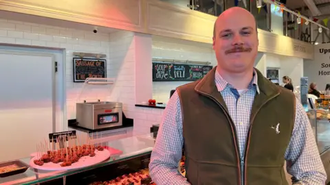 Matthew Slack stands in front of a butchers counter in a food hall. 