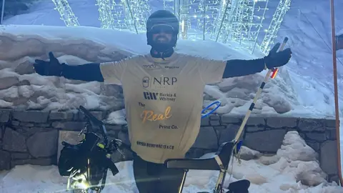 Mike Humphreys Mike Humphreys stood with his Chopper at Val Thorens in the French Alps. He is wearing a helmet and sun glasses and is standing in front of a wall and a curtain of fairy lights with his arms outstretched.

