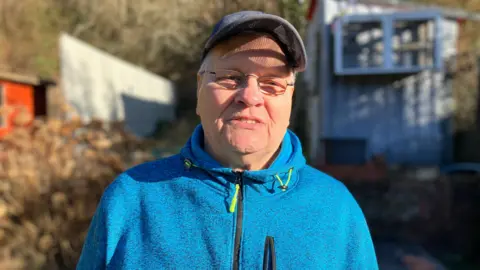 Brian Preece is wearing a blue hooded jumper and a baseball cap and is standing in his back garden, which backs on to the tip.
