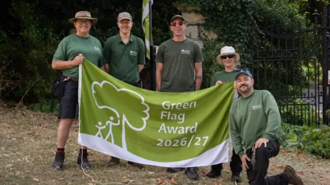 The University of Bristol Five people in green tops and dark trousers and shorts smile as they hold up a green and white flag saying "Green Flag Award 2026-27" in white letter