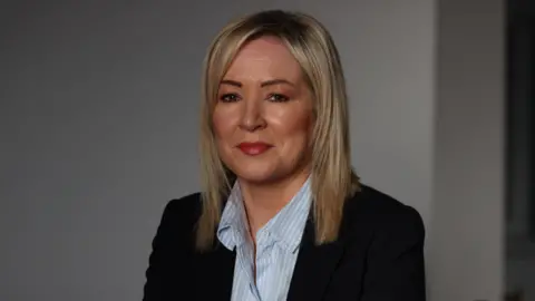 PA Media A close-up of Michelle O'Neill. She is a middle aged woman with shoulder-length blonde hair. She is smiling at the camera. She is wearing a black blazer and a blue and white striped shirt. She is sitting in front of a white wall.