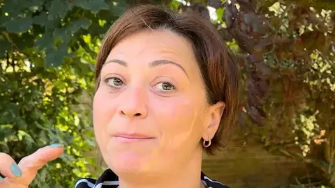 A woman with dark short hair in front of trees. She is looking to the camera