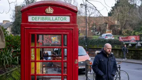 Getty Images A red traditional phone box, with a defibrillator sign on the top. There is a bright yellow defibrillator box inside. There is a man walking next to phone box, and cars are parked in the background