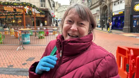 Marianne is wearing a red coat and blue gloves. She is smiling and standing in Birmingham's Christmas market. 