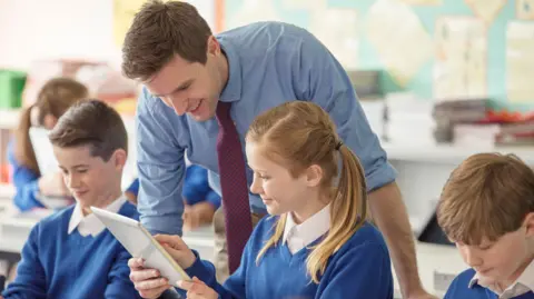 A stock image of a teacher in a classroom. He has short brown hair and is wearing a blue shirt with rolled up sleeves and a maroon and blue checked tie. He is leaning over to check the work of a girl aged about 11 who has blonde hair in pigtails and is pointing to something on an ipad. She has a blue jumper over a white shirt. Two boys with brown hair wearing the same uniform sit on either side of her. 
