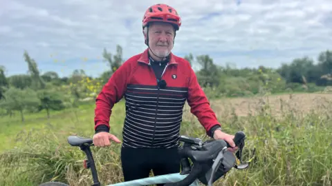 Peter Hargreaves is a man with grey hair and a grey beard. He is standing in front of a field, holding on to the handlebar and seat of his pale blue bicycle. He is wearing a red cycle helmet and a black, white and red cycling lycra top.