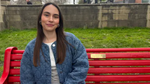 Faith has long brown hair and is smiling at the camera. She is sitting on a red bench with a gold plaque on it. She is wearing a white t-shirt and a blue jacket. Behind the bench is green grass and a stone wall.
