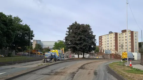 Park Lane in Gateshead, where demolition work on the Gateshead Flyover has been carried out. The road is closed and there are two vans and a number of heavy plant machinery, including a digger. There is metal fencing around the junction, with mounds of rubble and dirt around the area being worked on. In the distance there are two blocks of flats and another large building.