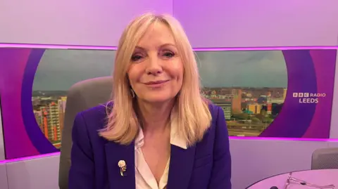 A woman with blonde hair, sitting in a radio studio. She is wearing a blue jacket and white shirt. There is a brooch on her jacket. The studio is lit in purple, and there is a skyline of Leeds behind her.
