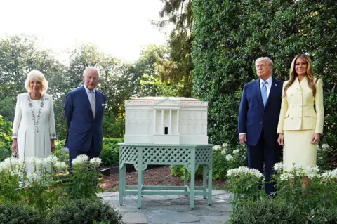Aaron Chown / Pool via Reuters King Charles III and Queen Camilla join US President Donald Trump and First Lady Melania, all dressed formally, in a green garden, beside a beehive in the shape of the White House