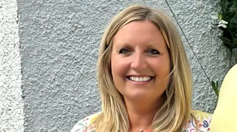 Head and shoulders shot of Nicola Purdie standing against a white wall. She has shoulder length blonde hair and wearing a flowery light coloured top. She is smiling directly at the camera.