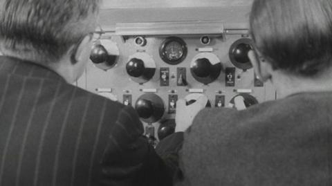 Two men sit at an audio mixing control panel.