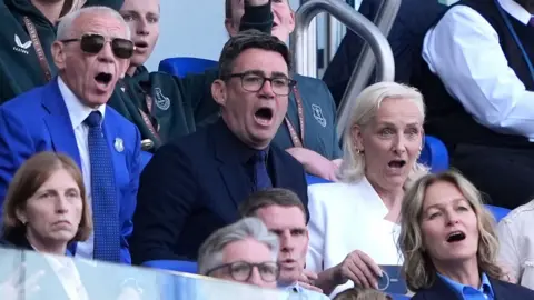 PA Media Andy Burnham is seated next to his wife Marie-France van Heel. They are sitting on blue seats, surrounded by people. Both of them have their mouths open in surprise while watching a football match.