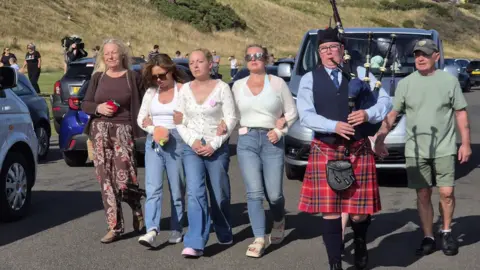 Five people with a piper in front walk in front of a grey private ambulance