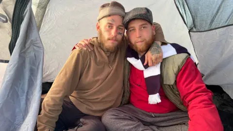 Two homeless men with ginger beards link arms inside of a tent wearing scarves.