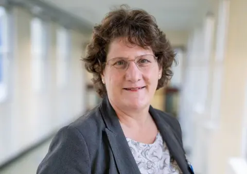 Queen Elizabeth Hospital Chief executive of the Queen Elizabeth Hospital in King's Lynn Alice Webster. She has short brown curly hair, wears frameless glasses, a grey blazer and pink top patterned with grey swirls. 