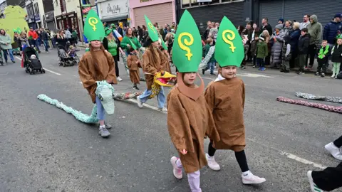 Pacemaker A group of children participating in the St Patrick's Day parade in Downpatrick. They are wearing brown robes and large green St Patrick hats. Two of them are carrying large fake snakes.