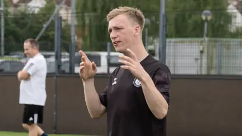 The FA A man is wearing a black top and is coaching people on a football pitch.