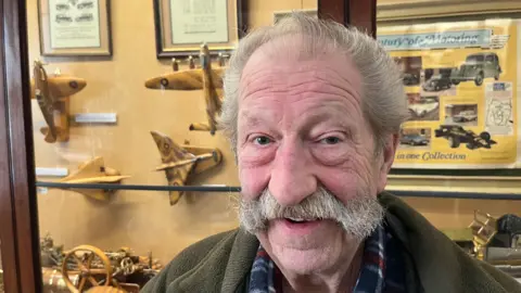 Fred Clarke, 82, has grey hair swept back over his head and a large handlebar moustache he is wearing a brown fleece with checked shirt underneath. Behind him is a display case containing some of his models including a traction engine, spitfire aircraft, and more, all made of wood and brass and highly detailed.