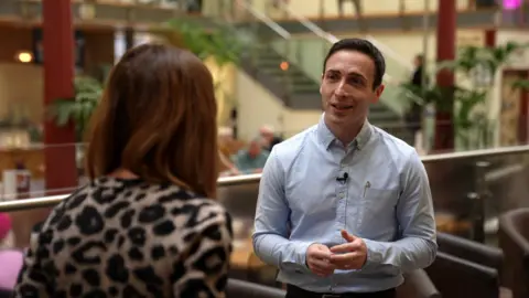 Dan Westgarth stands in a open atrium, with large plants in the background. He has dark hair and wears a blue shirt.