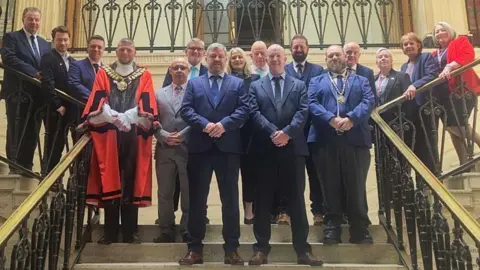Barnsley Council A large group of people posed on a grand staircase inside Barnsley Town Hall. The group is arranged on several levels of the stairs, with some people standing at the front and others positioned higher up on the steps and landings.
Above the group, a sign reads "Council Chamber".