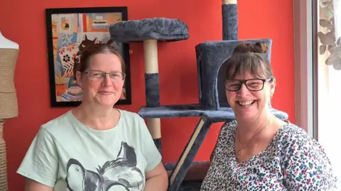 Two middle-aged women smile at the camera while standing in front of a grey cat tower. The wall behind them is red and has an artwork featuring a cat on it.