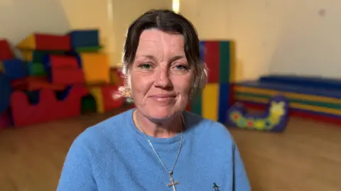 Tanya smiling into the camera. She has short dark hair and is wearing a light blue jumper with a silver chain and cross around her neck. She's pictured in a playroom setting with soft toys in the background.   