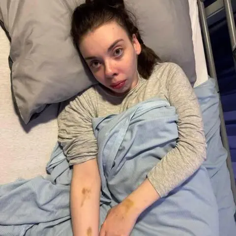 Katie Robertson A woman laying in a hospital bed wearing a grey long sleeved jumper.