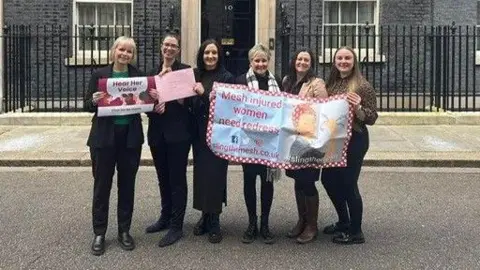 Susan McLarnon Six women, all wearing black, standing in front of Downing Street. They are holding posters reading 'mesh injured women need redress'. 